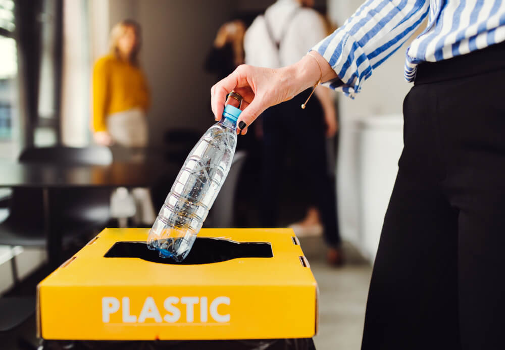 A person in a striped shirt places a plastic bottle into a yellow bin labeled "PLASTIC." The background shows a blurred office environment with people.
