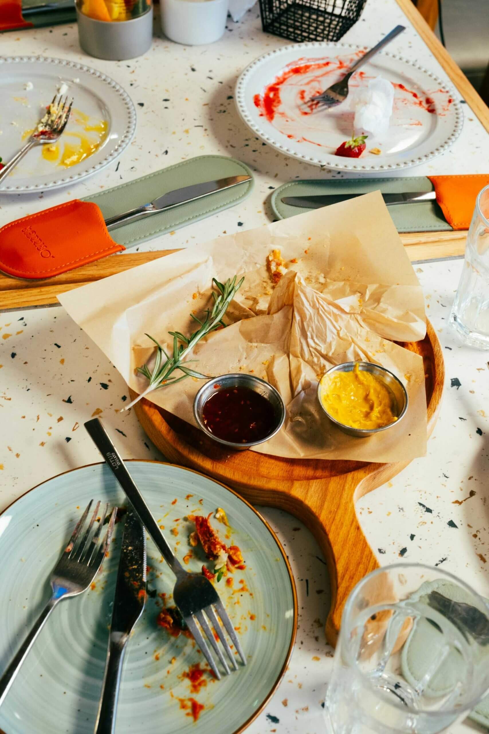 Restaurant table after a meal, with empty plates, a wooden serving board with parchment paper, a rosemary sprig and two dipping sauces